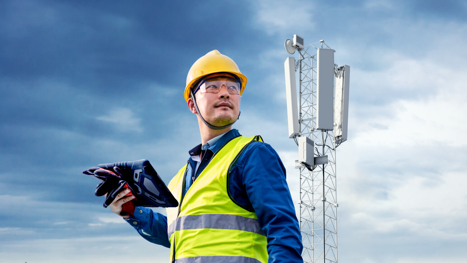Engineer with tablet near telecom tower under cloudy sky.