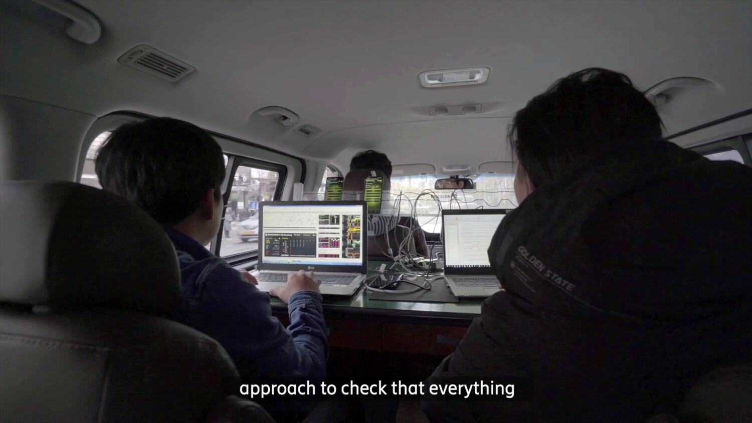Three people in vehicle working on laptops with cables.
