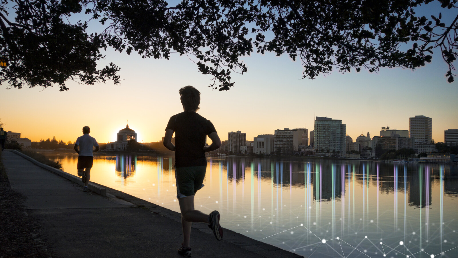 Two people jogging by lake at sunrise, city skyline behind.