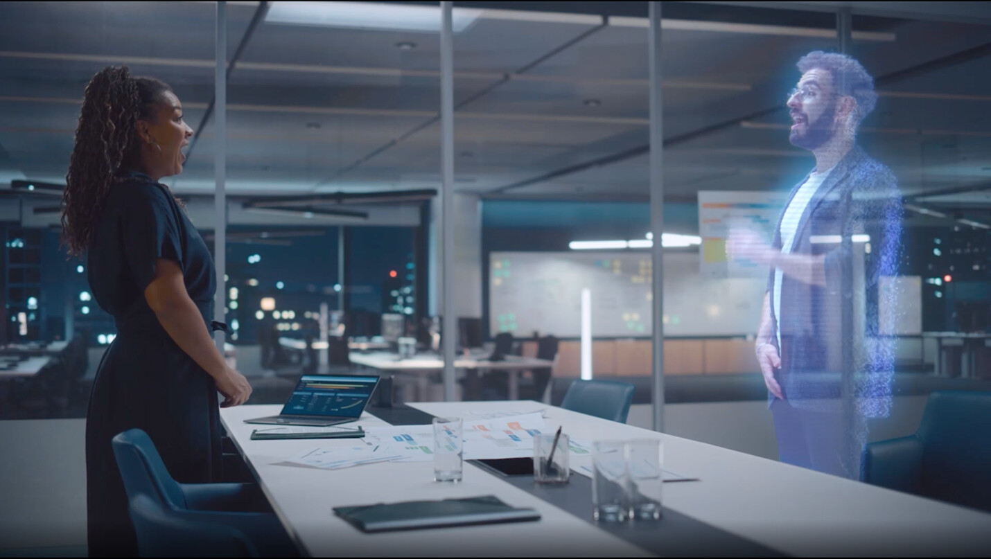 Person in office with holographic colleague and laptop.