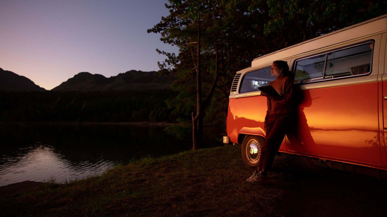 A woman with a tablet beside a camper van in the evening.