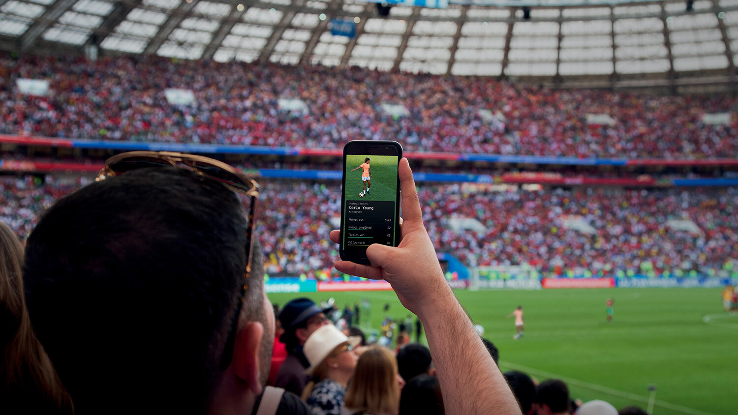Man uses smartphone with stats overlay at crowded sports stadium.