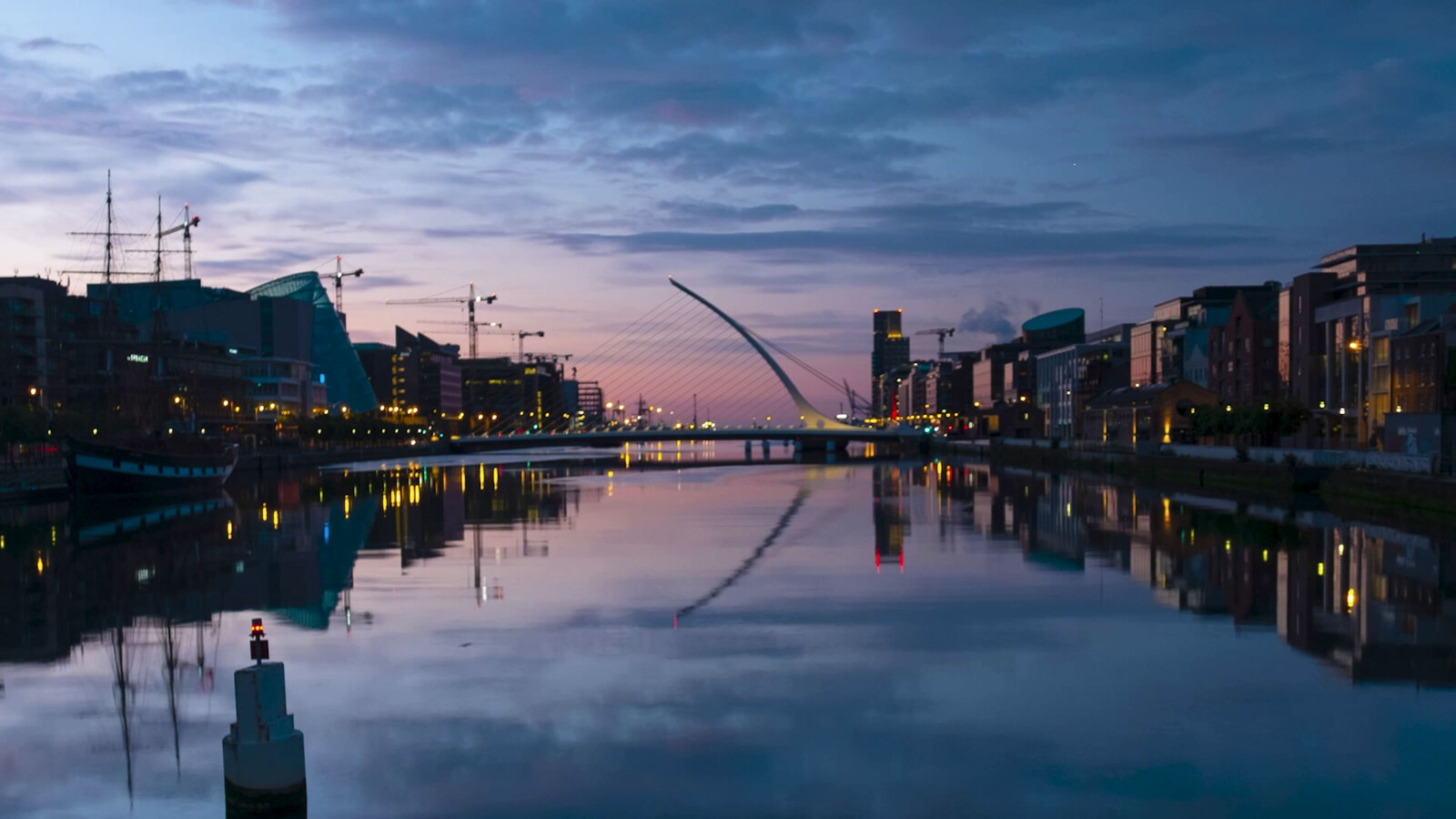Twilight cityscape with a modern cable-stayed bridge and city lights reflecting on calm river water.