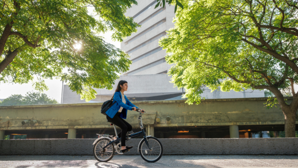 Woman in a blue jacket rides a bike on a tree-lined path with a modern building in the background.
