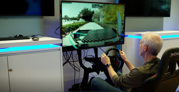 A person using a driving simulator setup with a steering wheel and pedals, controlling a remote vehicle displayed on a large screen.