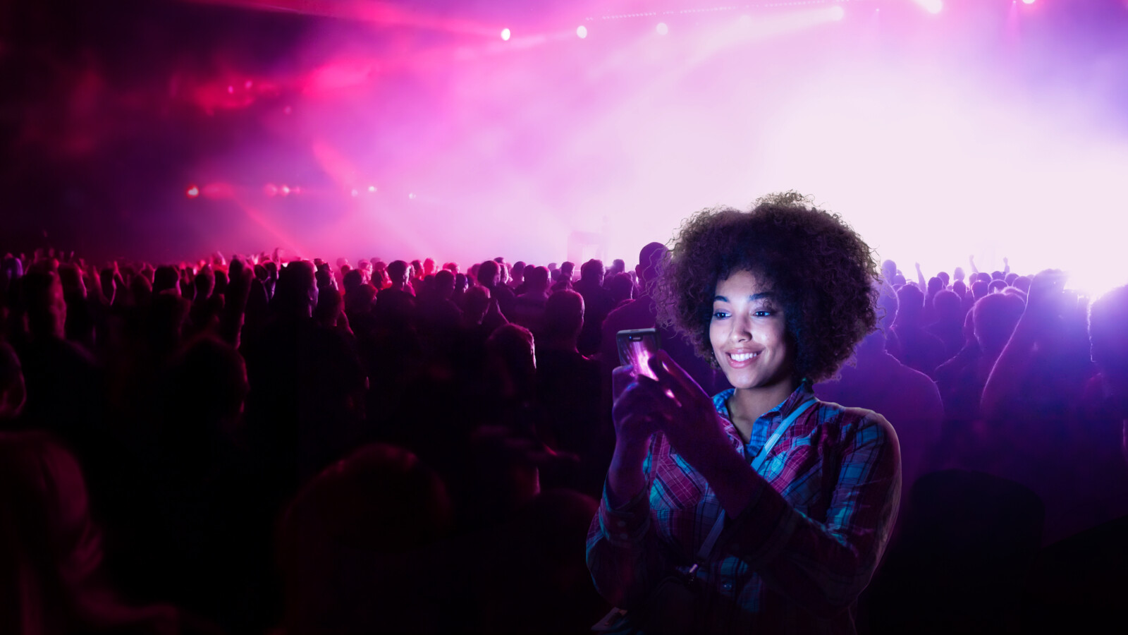 Crowd at concert, person filming with phone, bright lights.