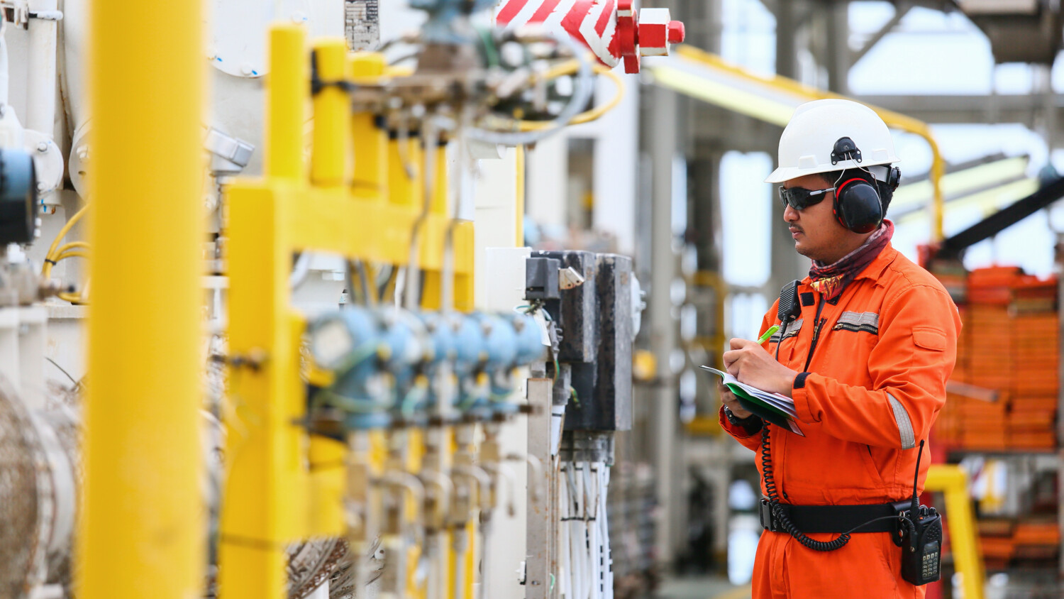Worker inspects gauges in industrial setting with clipboard.