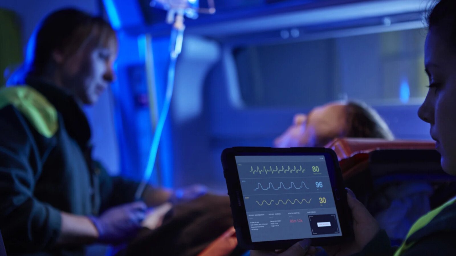 Paramedics monitor patient in ambulance using a tablet.