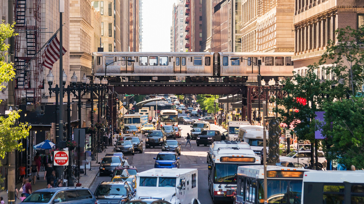 Busy city street with traffic, train overhead, and crowds.