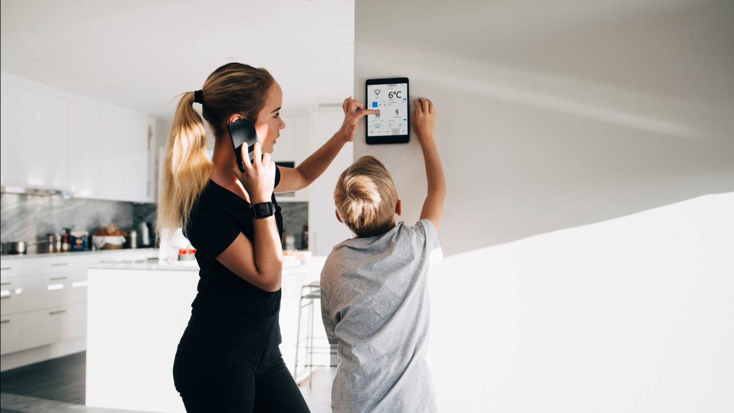 Two people use smart home wall device in kitchen.