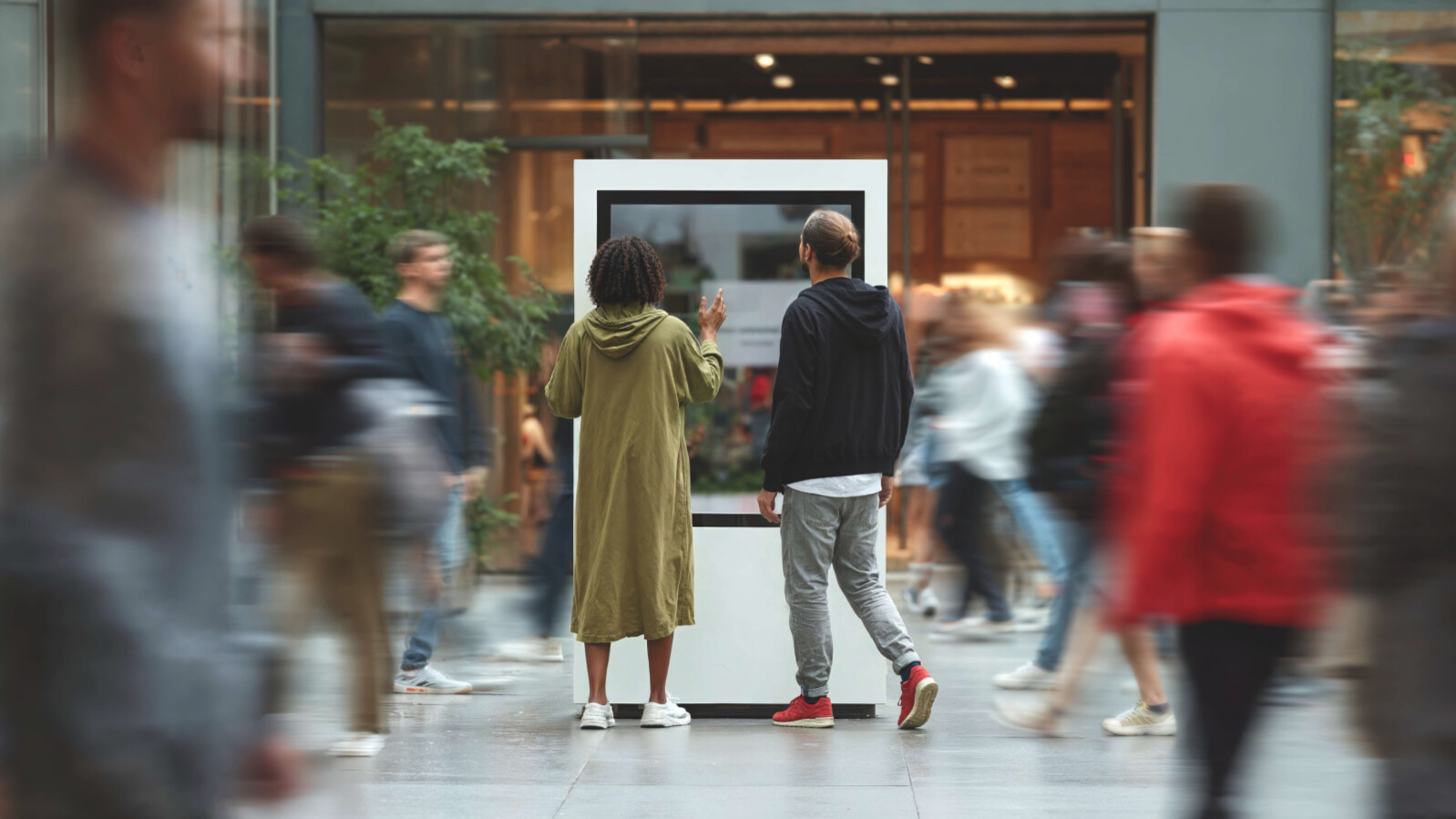 Two people interacting with digital display kiosk in busy public space with motion blur crowd