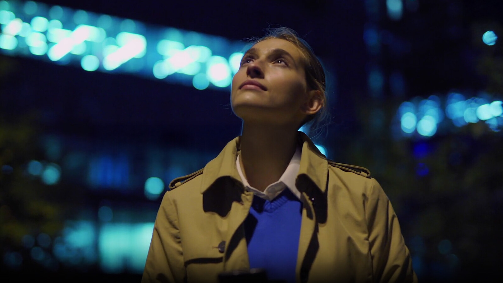 Woman in trench coat looks up at night with city lights in background.