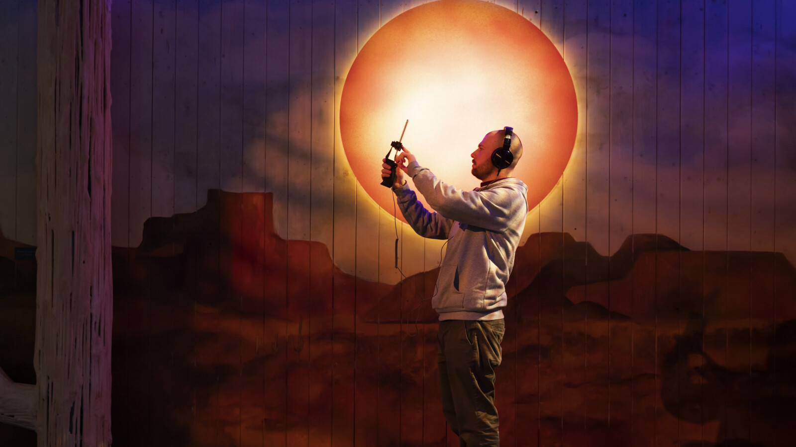 Man with headphones holding up a mobile phone in a room with orange light.