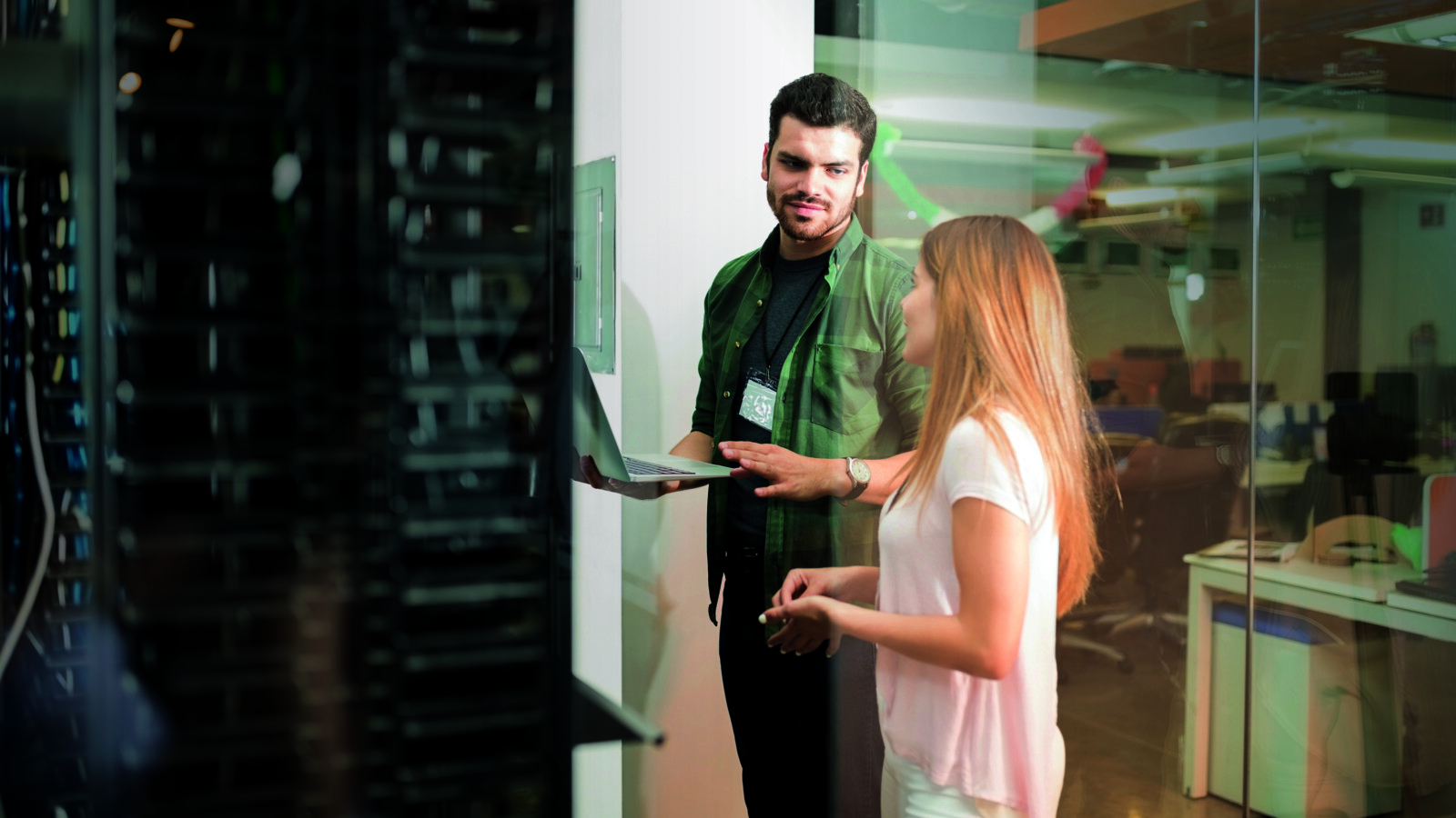 Two people by a server rack, one with laptop, in office setting.