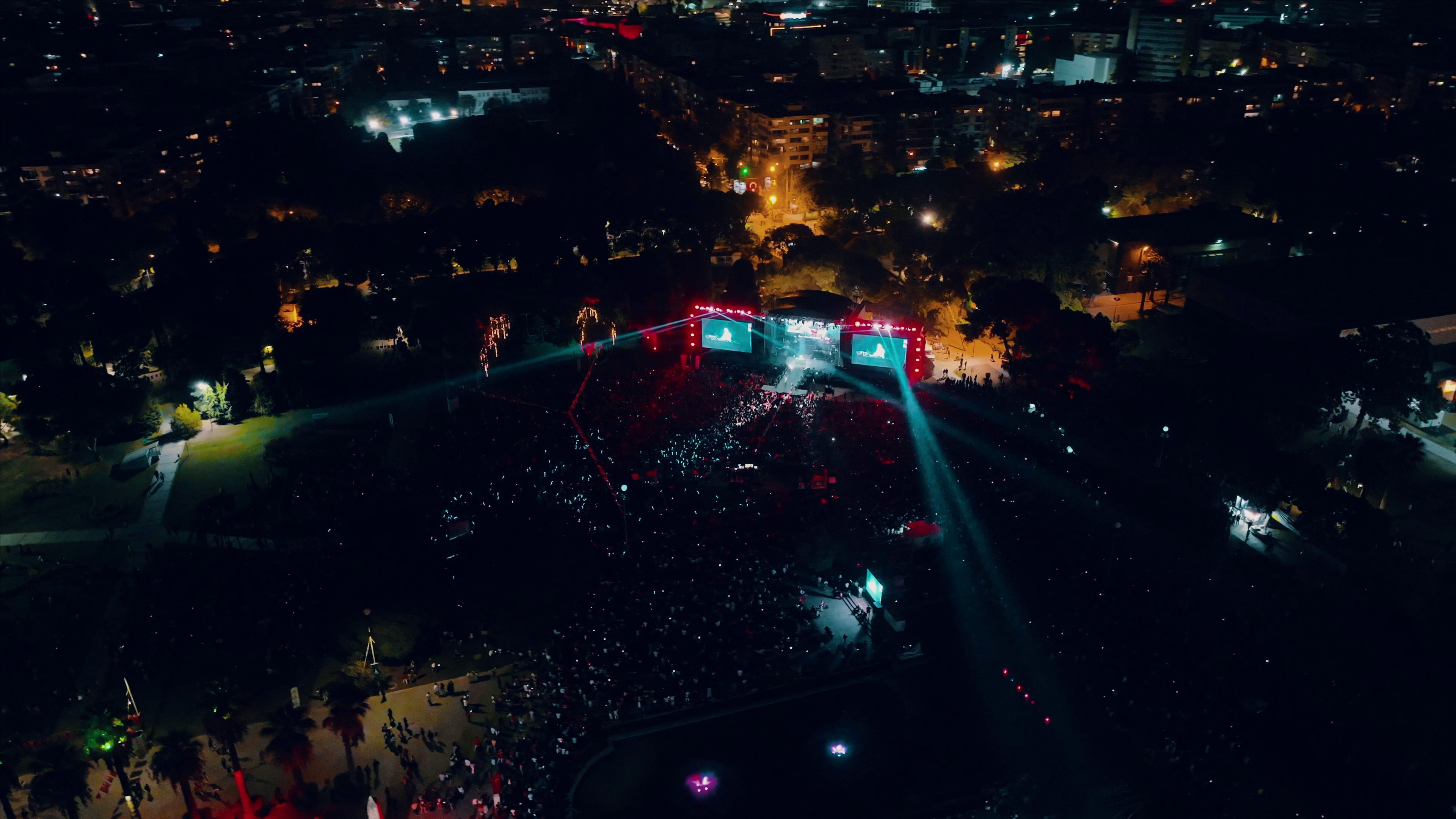 Aerial view of a night concert with stage, crowd, and city lights.