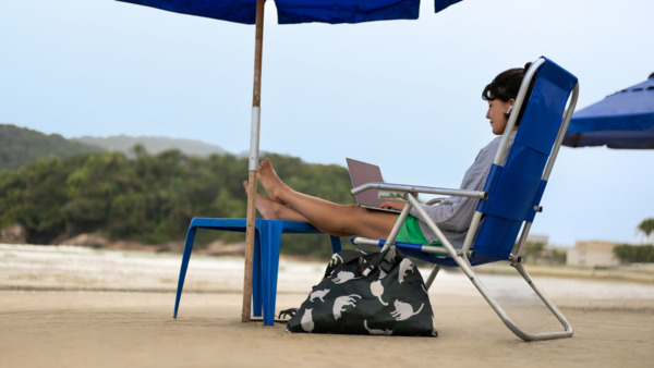 Person on beach chair with laptop under blue umbrella.
