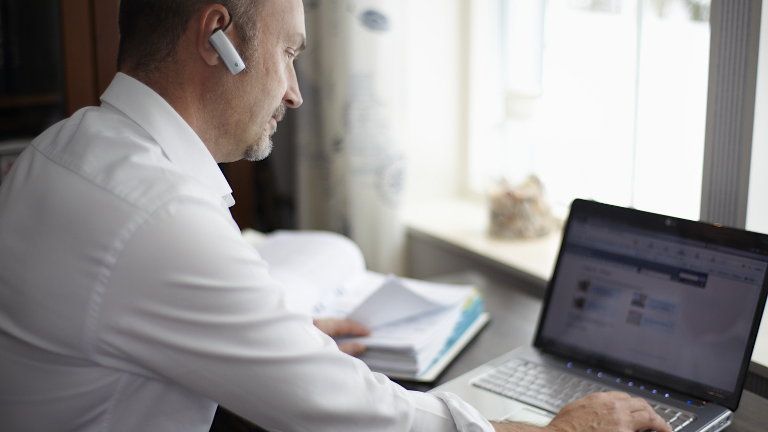 Man wearing bluetooth ear piece and working at a laptop computer.