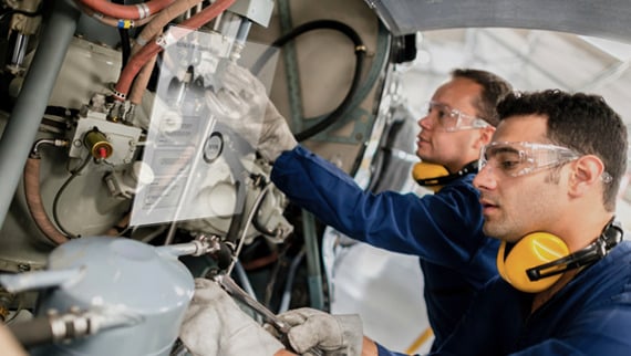 Two workers in gear fix machinery with cables and parts.