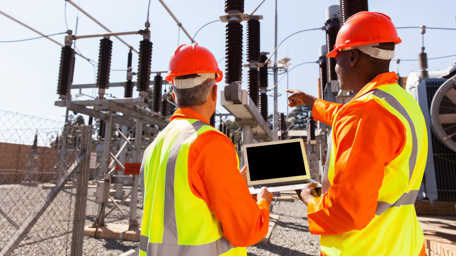 Engineers with laptop at outdoor electrical substation with transformers. 