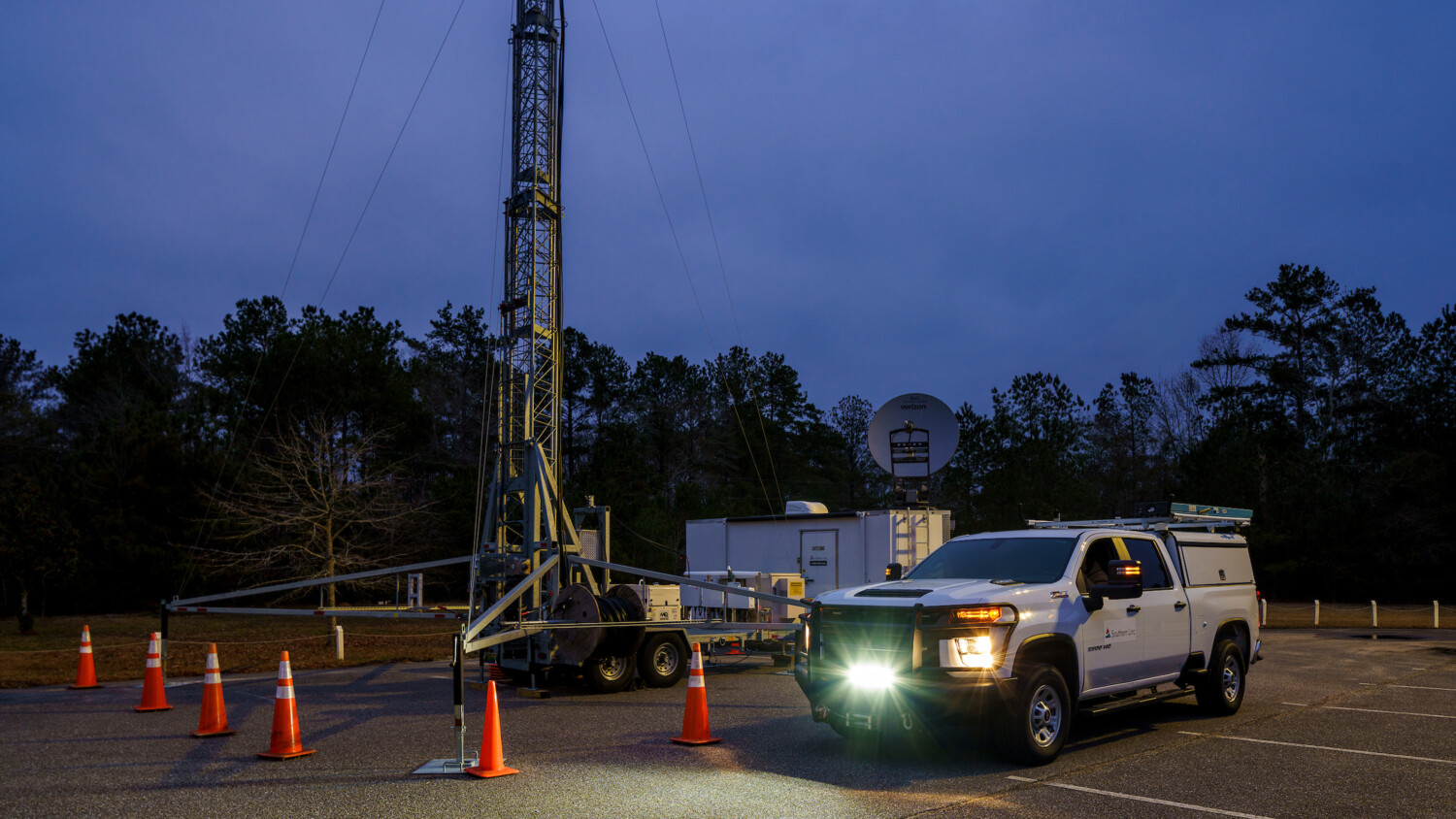 Utility truck by tower and trailer at twilight with cones.