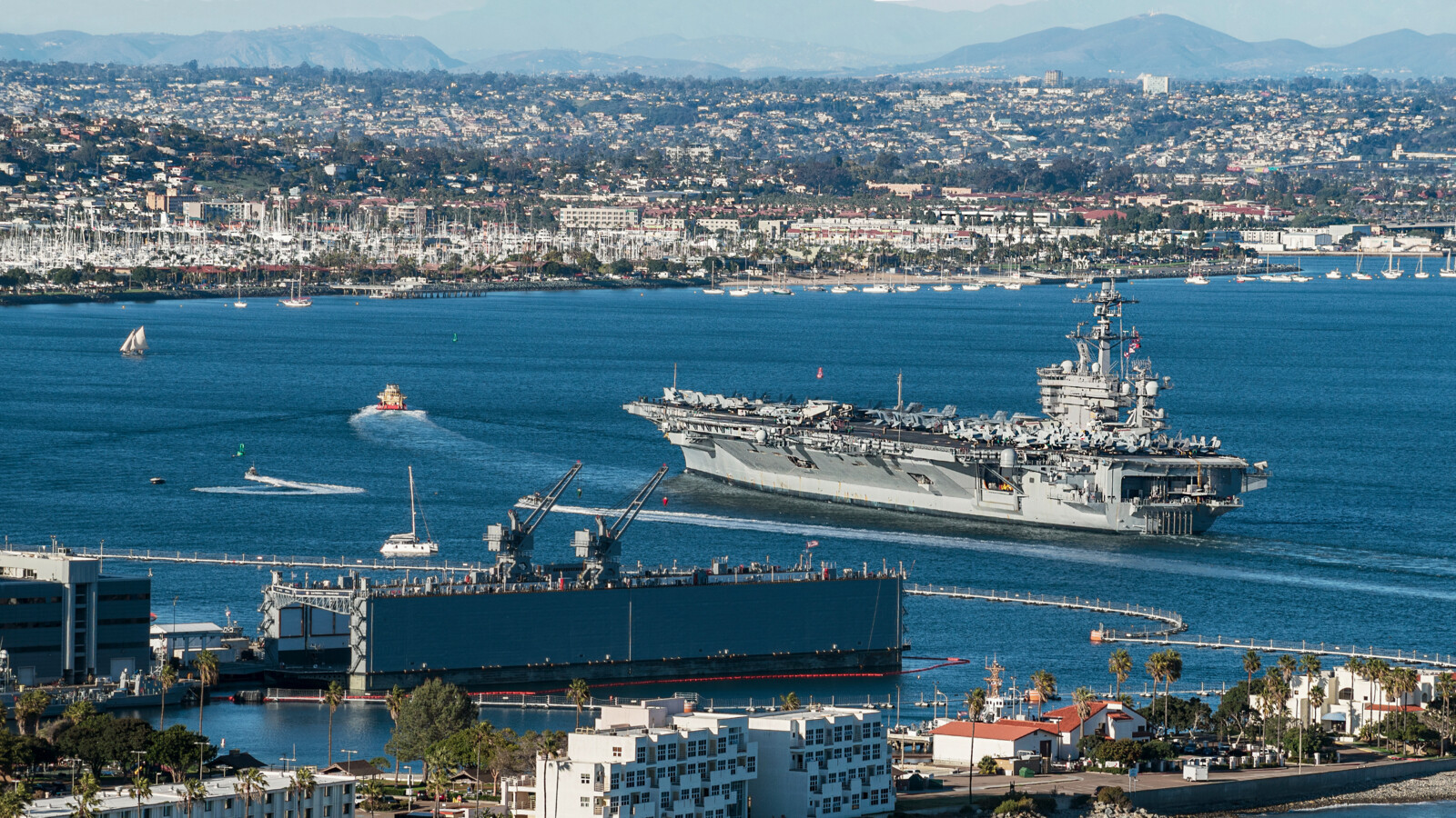 Aircraft carrier in harbor with city, boats, and mountains behind.