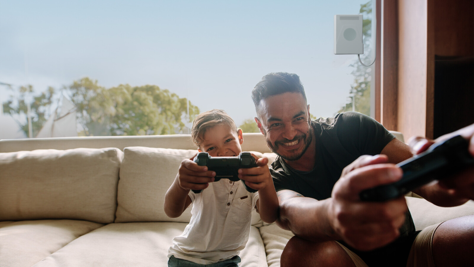 Father and son gaming gaming on couch with trees outside window.