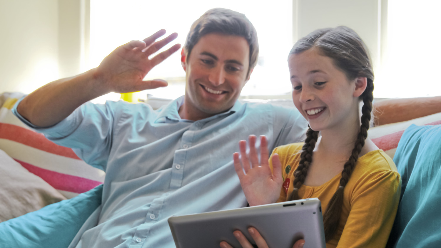 Un padre y una hija están usando una tableta y agitando durante una videollamada.