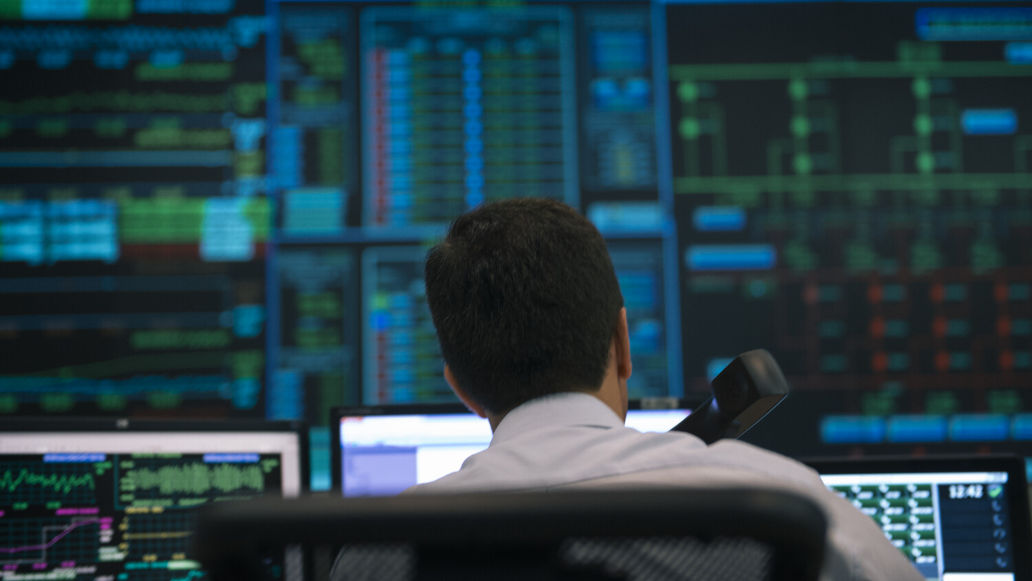 Man sits in a control room with multiple screens displaying data, phone in hand.