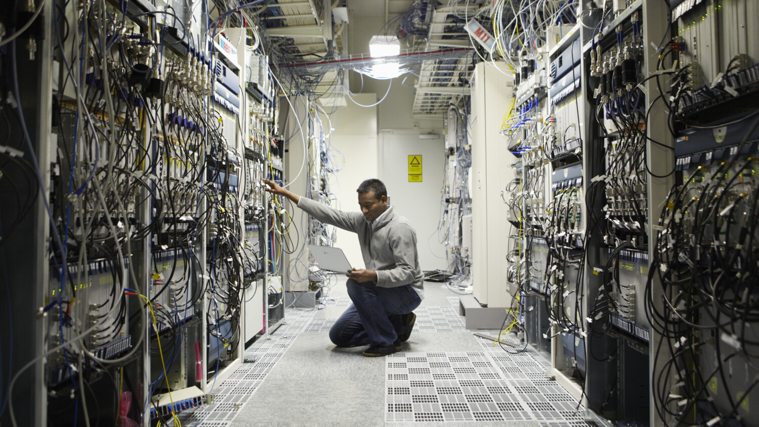 Technician works on laptop in server room with cables.
