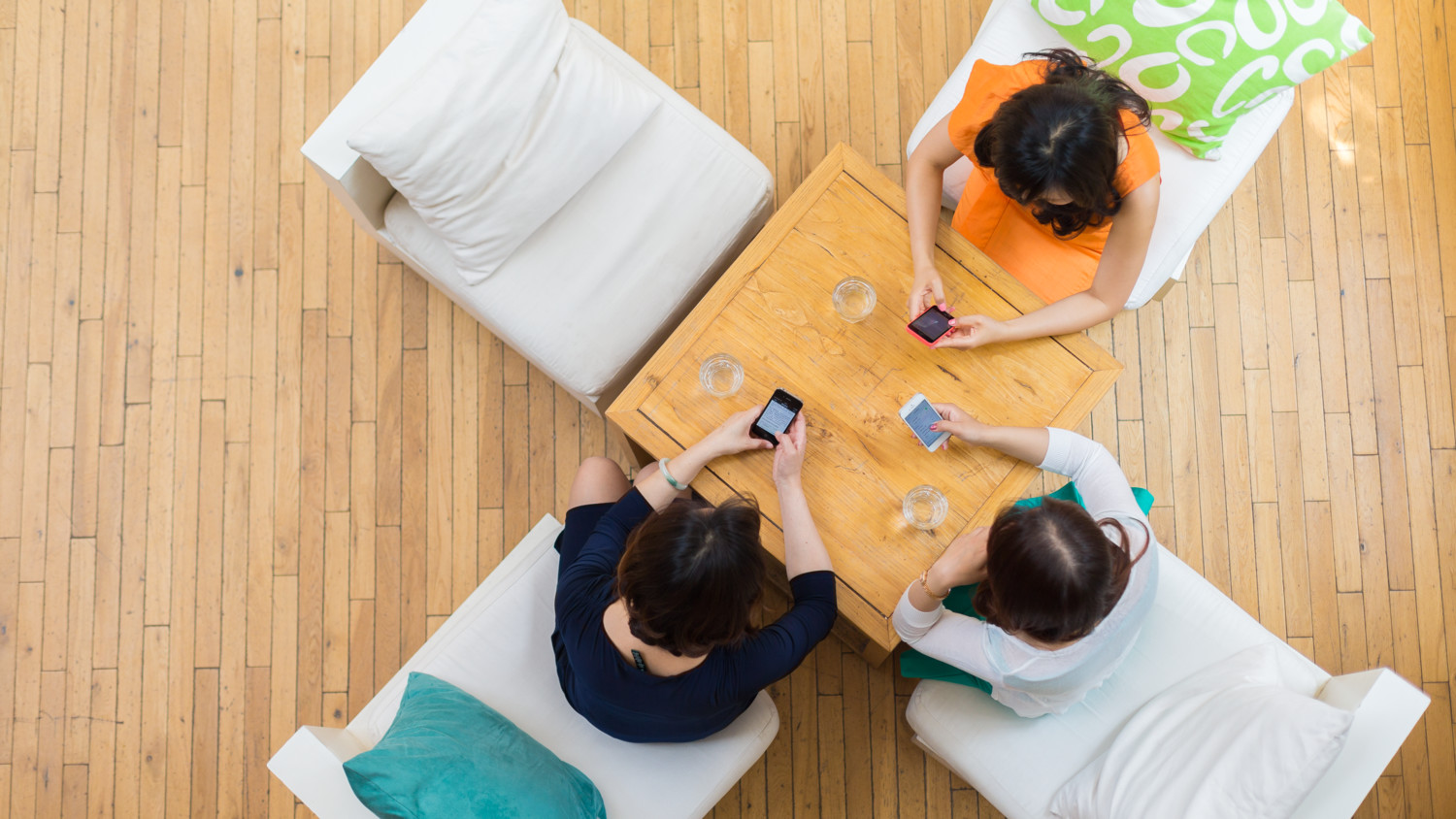 Três mulheres sentadas ao redor de uma mesa, usando seus telefones.