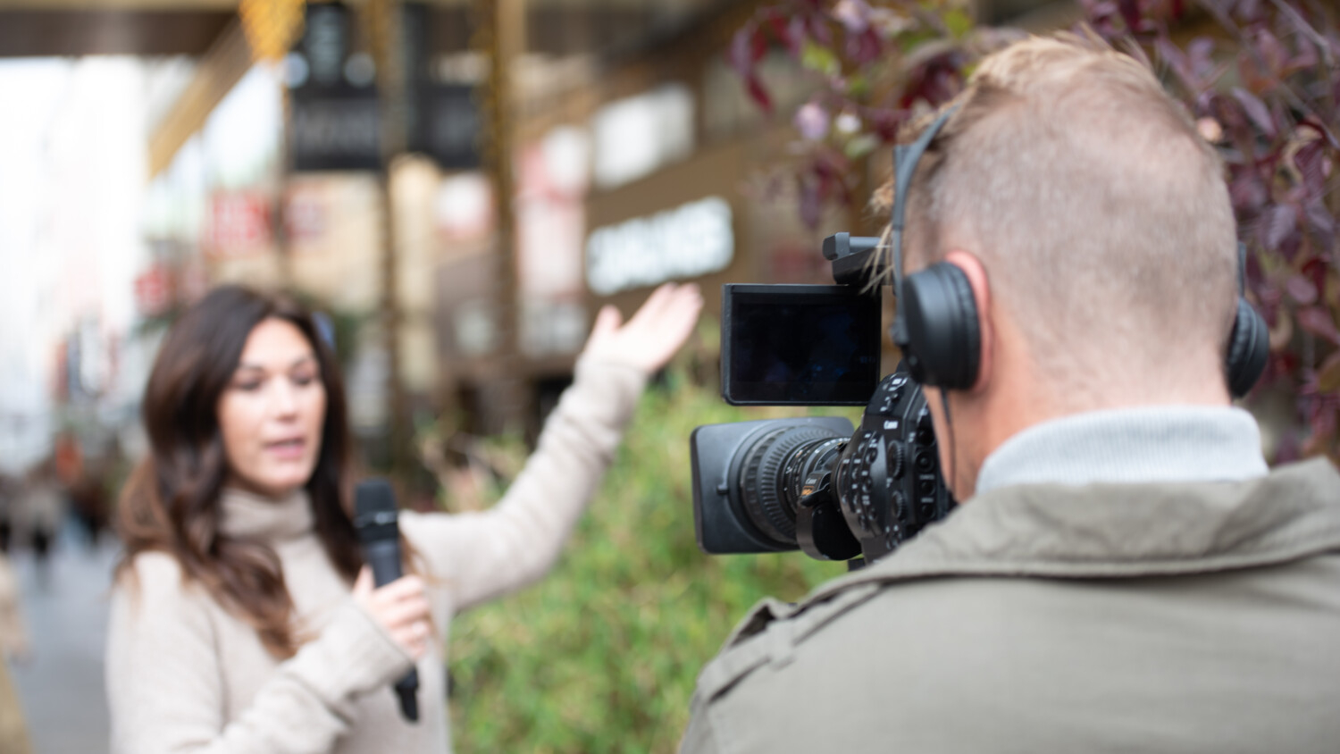 Camera operator filming presenter with microphone outdoors.