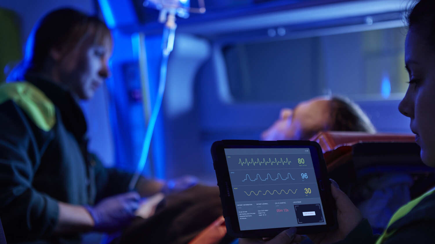 Paramedics monitor patient in ambulance using a tablet.