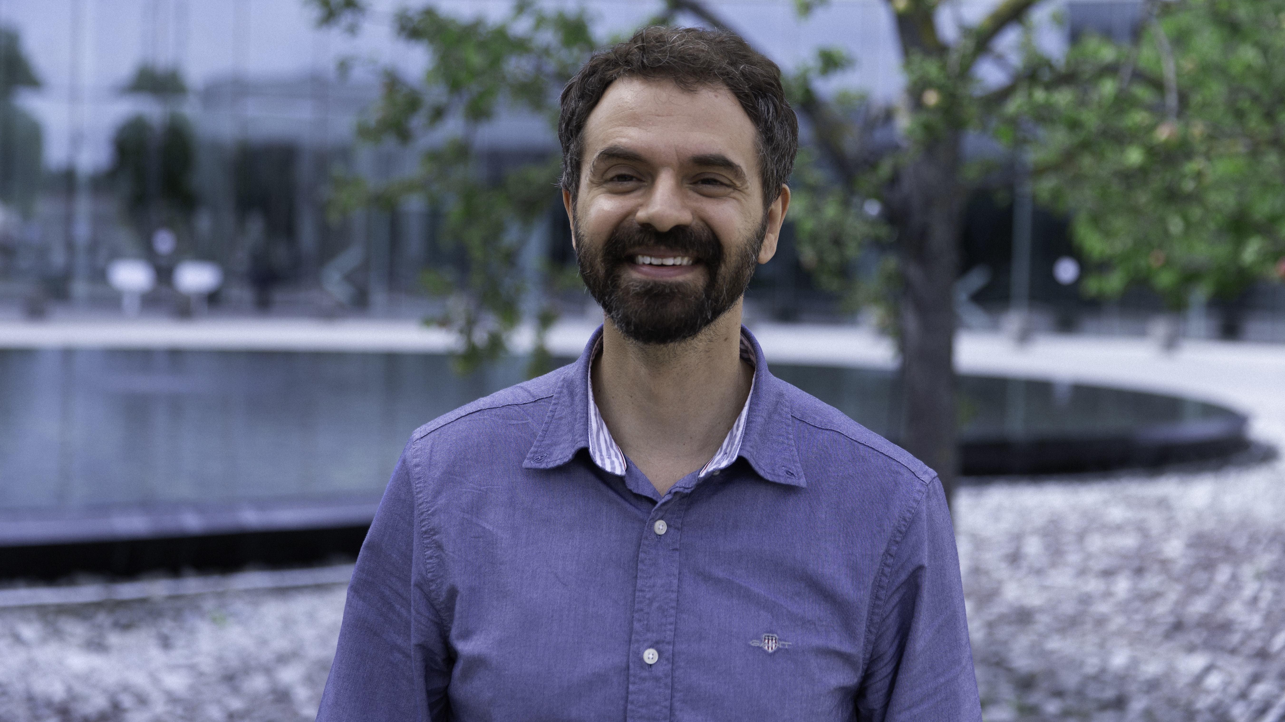 Konstantinos Vandikas the winner of the inventor of the year award standing outside Ericsson's headquarters.  He has a beard and is smiling while wearing a blue shirt. 