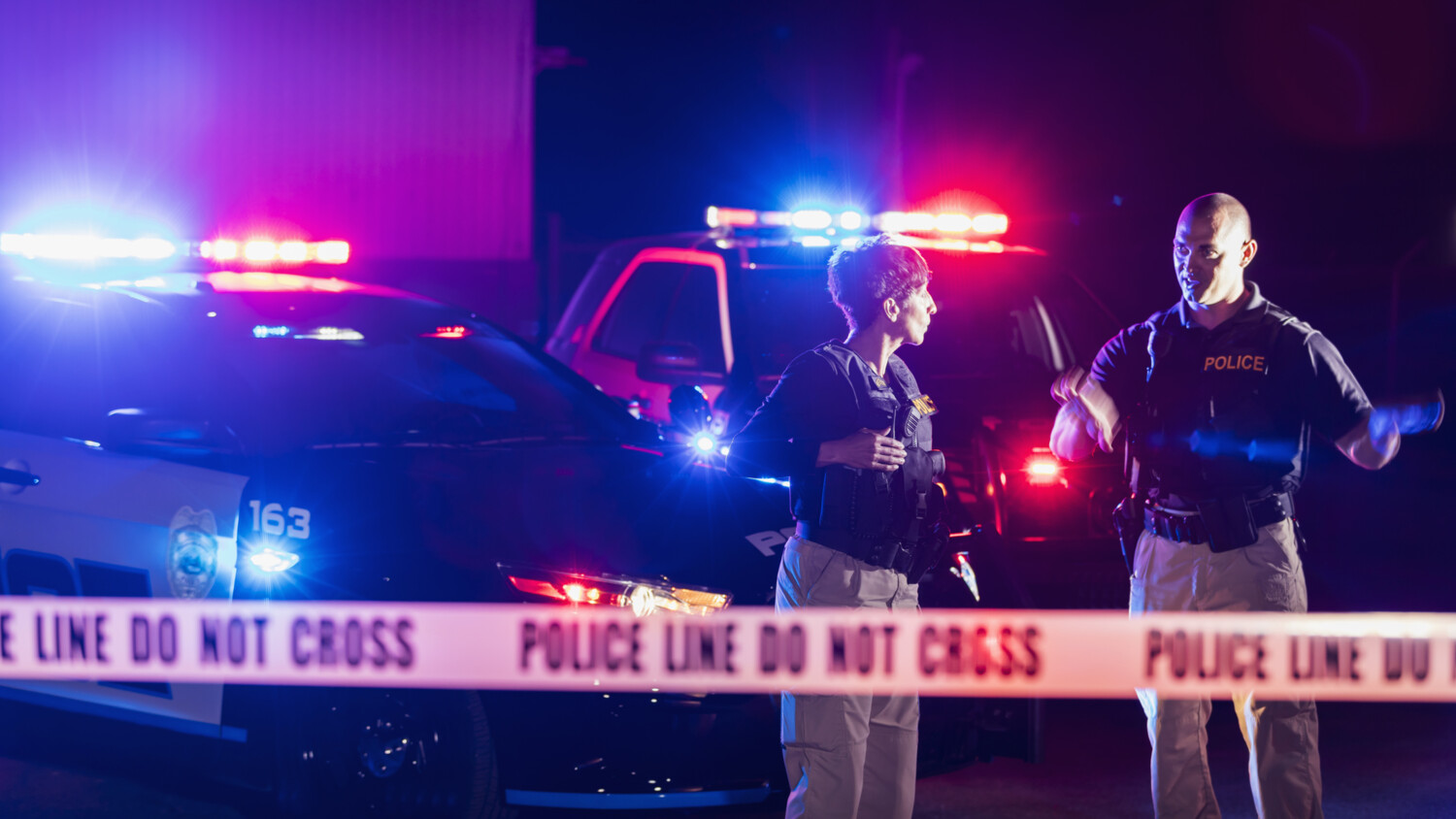 Police officers at night behind tape, patrol cars flashing red and blue lights.