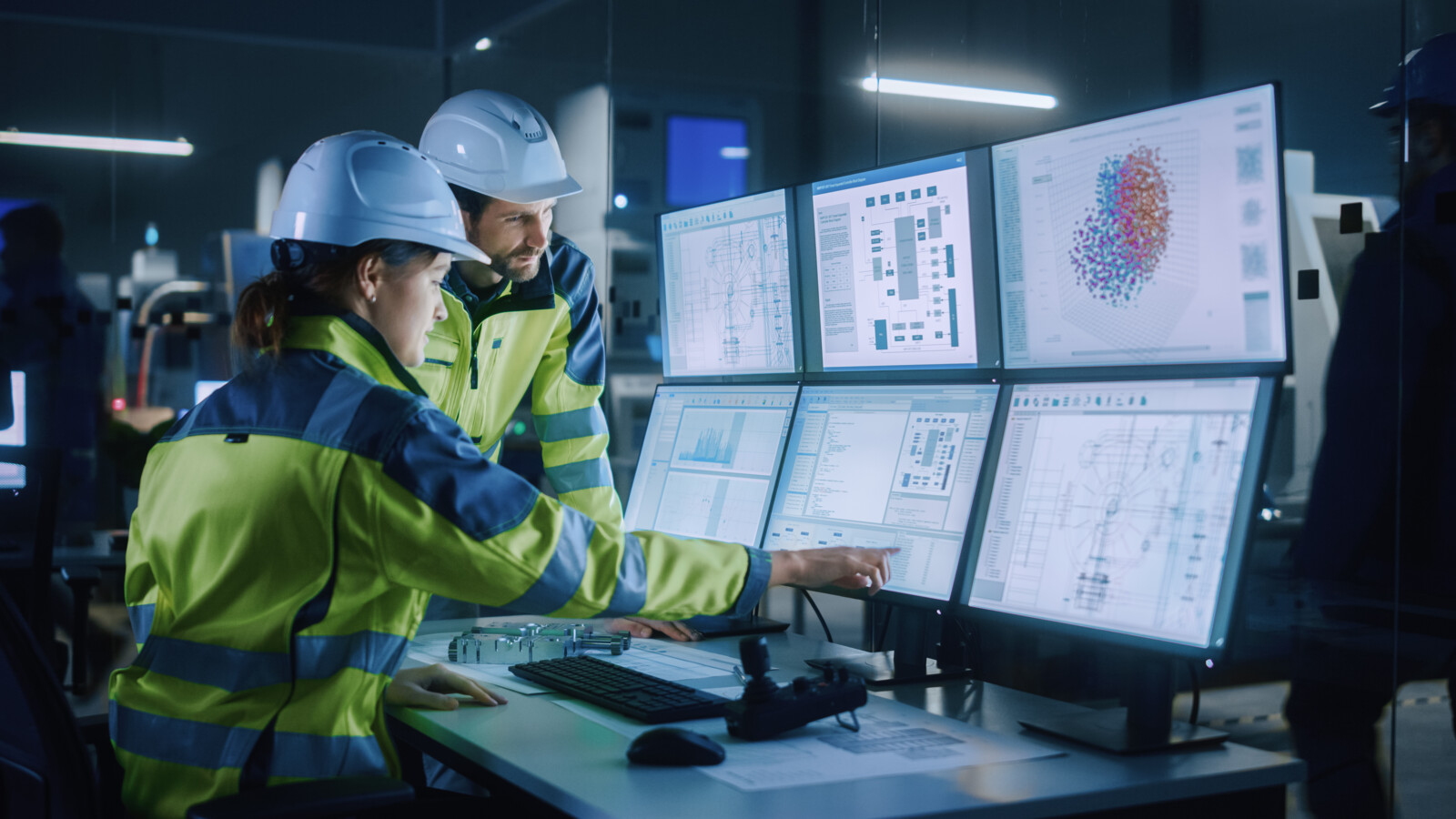 Two workers in control room reviewing data on multiple computer screens.